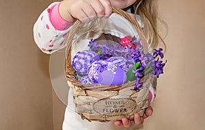 A girl holding a basket with painted eggs and flowers in lilac flowers, for Easter.