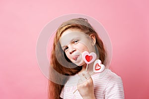 Girl holding big heart-shaped lollipops in her hands on a pink background.