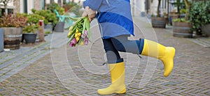 The girl is holding a bouquet of fresh tulips in her hands, yellow and pink in a blue cloak and yellow rubber boots. in a city on