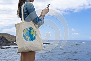 Girl holding cotton canvas while typing with her cellphone in the sea. Conscious Woman using ecology carrier handbag and messaging