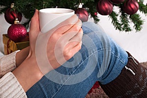 Girl holding cup of coffee in hand at legs with warmers and Christmas tree in the background