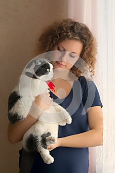 A girl is holding a fluffy cat in a scarf that looking out of the window
