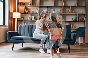 Girl holding gift box behind her. Happy holidays. Young mother with her daughter is together at apartment