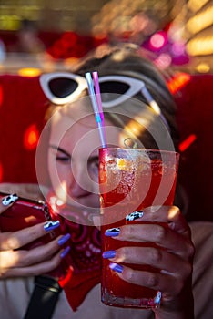 The girl is holding a glass with a bright cocktail and a red phone. The sun shines through the window bars