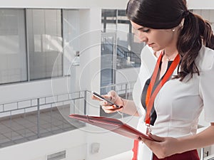 The girl is holding the phone in her hands. Beautiful Smiling Businesswoman Standing Against White Offices Background. Portrait of