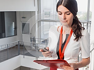 The girl is holding the phone in her hands. Beautiful Smiling Businesswoman Standing Against White Offices Background. Portrait of