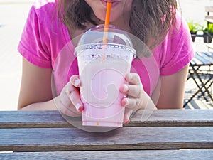 Girl holding plastic glass of strawberry milkshake with straw in cafe on summer day