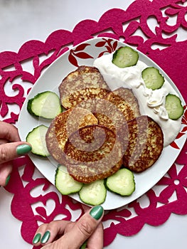 The girl is holding a plate with potato pancakes, sour cream and fresh cucumber, sliced â€‹â€‹with ringlets.