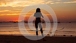 Girl holding smart phone and taking red sunset photo on the tropical beach.  Silhouette of a woman, yachts and mountains on