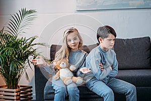 Girl holding teddy bear while offended boy sitting on sofa at home