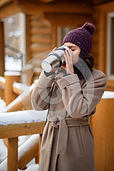 A girl holding a thermos with a hot drink. A mug of coffee in her hands. Winter alley in the city, a young girl walks