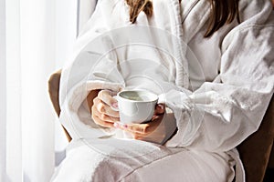 Girl holding a white coffee mug. In a white coat. Coffee in bed. Morning begins with coffee. Coffee in the hands of the bride