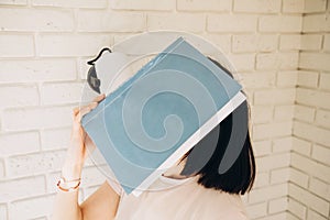 The girl holds a big blue book in her hand and covers her face against the background of a white brick wall