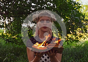 Girl holds a burning book in her hands. A young woman in a forest burns a book