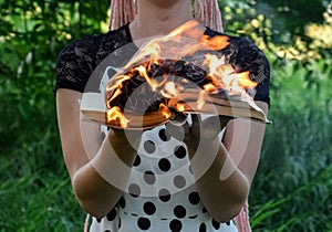 Girl holds a burning book in her hands. A young woman in a forest burns a book