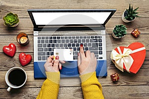 Girl holds debit card, laptop, chooses gifts, makes purchases, cup of coffee, two hearts, succulents on wooden table