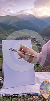 A girl holds in her hand a paintbrush near a white canvas on the background of a mountain settlement on a summer evening