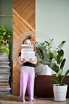 A girl holds a large stack of books against the background of a home.