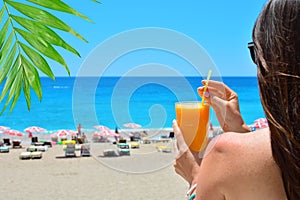 Girl holds orange fresh juice in her hand. Blue sea and beach on a backdrop