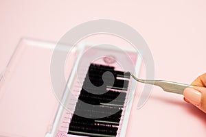 Girl holds tweezers over a pink box with black eyelashes on a pastel background. Eyelash Extension tools
