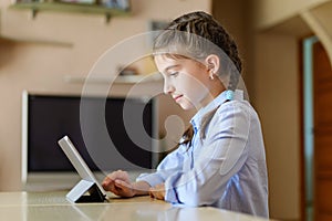 The girl at home sits at a table and uses a tablet.