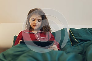 Girl in homemade pyjamas sitting in bed looking at tablet, lifestyle.