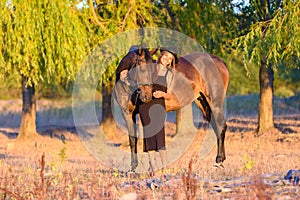 A girl with a horse stands against the background of trees, the rays of the setting sun fall on them