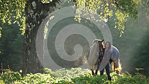 Girl with a horse on a walk in the woods early in the morning next to a beautiful tree in the rays of dawn