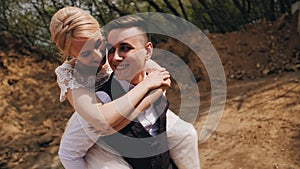 Girl hugs her boyfriend from behind. She presses her cheek to him and smiles. The guy smiles back. Young beautiful couple in natur