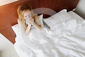 Girl is ill, sneezes in white bed with napkin at home sunny day