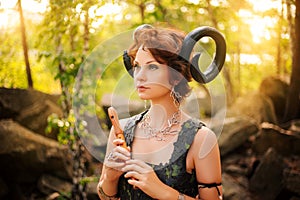 A girl in the image of a faun stands with a wooden flute. Model with horns on his head and with furry pants in the woods among the