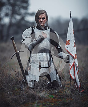 Girl in image of Jeanne d`Arc in armor kneels with flag in her hands and sword on meadow. photo