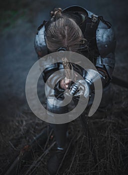 Girl in image of Jeanne d`Arc in armor and with sword in her hands kneels against background of dry grass.