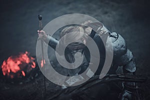 Girl in image of Jeanne d`Arc in armor and with sword in her hands kneels against background of fire and smoke.