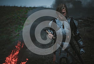 Girl in image of Jeanne d`Arc in armor and with sword in her hands stands against background of fire and smoke.