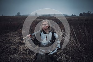Girl in image of Jeanne d`Arc in armor and with sword in her hands stands on meadow. Closeup.