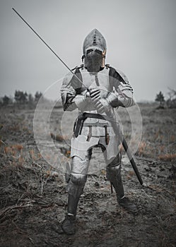 Girl in image of Jeanne d`Arc goes on meadow in armor and helmet with closed visor and sword in her hands.