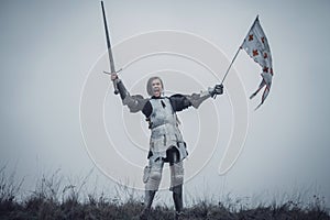 Girl in image of Jeanne d`Arc stands in armor and issues battle cry with sword raised up and flag in her hands.