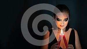 Girl in image of witch looks straight into frame with burning candle in hands on black background. Concept of Halloween