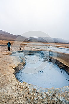 Girl infront of bubbling hot mud pool with mountains and s-shaped road in the background in the Hverarond volcanic area in Iceland