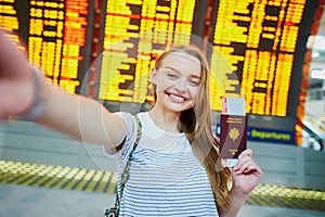 Girl in international airport, taking funny selfie with passport and boarding pass near flight information board