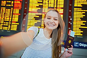 Girl in international airport, taking funny selfie with passport and boarding pass near flight information board