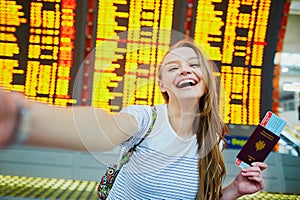 Girl in international airport, taking funny selfie with passport and boarding pass near flight information board