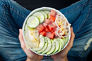 Girl in jeans holding hawaiian watermelon poke bowl with avocado, cucumber, mung bean sprouts and pickled ginger. Top view