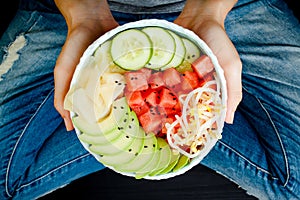 Girl in jeans holding hawaiian watermelon poke bowl with avocado, cucumber, mung bean sprouts and pickled ginger. Top view