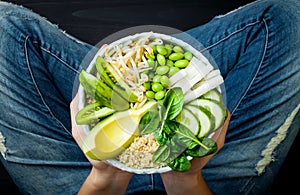 Girl in jeans holding vegan, detox green Buddha bowl with quinoa, avocado, cucumber, spinach, tomatoes, mung bean sprouts, edamame