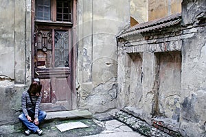 A girl in jeans, a striped sweater and sneakers sits on the doorstep by the wooden door of a very old house