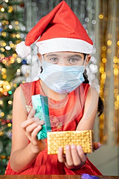 Girl kid in medical mask with Christmas hat and decorated background opening gift box - concept of Gift sharing, distant