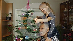 Girl kid with senior grandpa decorating artificial Christmas tree with ornaments and toys at home