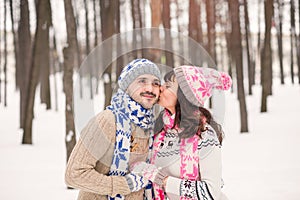 Girl kissing her boyfriend on the cheek in winter outdoors. Wearing cozy warm clothes, knitted hat and gloves . Winter dating con